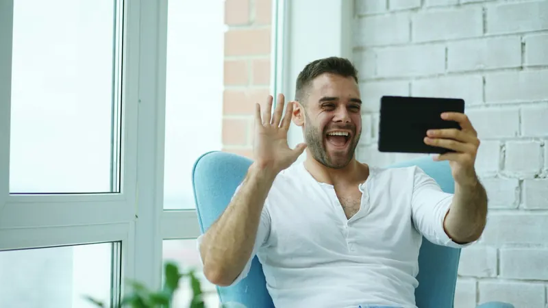 Un homme salue de la main pendant un appel vidéo sur sa tablette, assis dans un fauteuil